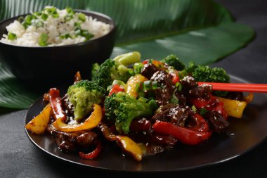 Masada kırmızı ve sarı dolmalık biberli Asya teriyaki bifteği brokoli ve susam tohumları var. Baharatlı teriyaki bifteği. Arka planda sebze ve pirinçle kızartılmış.