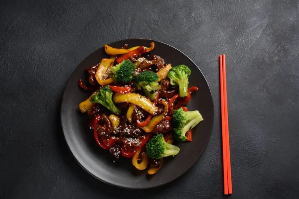 Masada kırmızı ve sarı dolmalık biberli Asya teriyaki bifteği brokoli ve susam tohumları var. Baharatlı teriyaki bifteği. Arka planda sebze ve pirinçle kızartılmış.