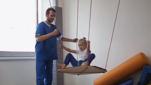 Kind meisje met fysiotherapeut swingen op swing tijdens zintuiglijke integratie sessie. Ontwikkelingsfuncties van het vestibulair apparaat bij kinderen. — Stockvideo