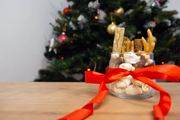 Натюрморт собачьей еды и здоровые угощения с красной ленточкой на фоне рождественской елки с копировальным пространством. рождественский подарок для домашних животных. — стоковое фото