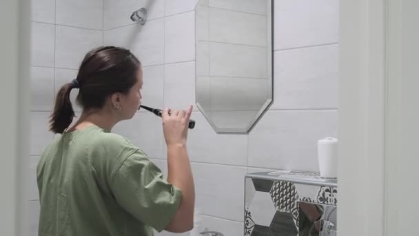 Женщина чистит зубы электрической зубной щеткой в домашней ванной. Ошибка в зеркале — стоковое видео