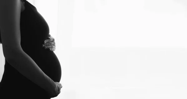 Silhouette di donna incinta in abito a casa dalla finestra toccare pancia. Foto in bianco e nero. — Foto Stock