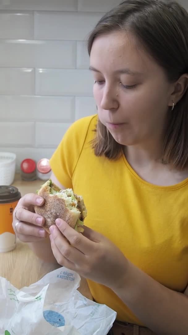 O tânără care mănâncă un hamburger delicios acasă. Conceptul de livrare a alimentelor în timpul carantinei. Supraalimentarea și stresul în singurătate și izolare. — Videoclip de stoc