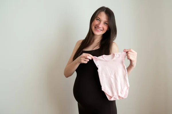 幸せな若い妊婦は新生児の女の子のための服アイテムのボディスーツを準備します。幸福妊娠の概念 — ストック写真