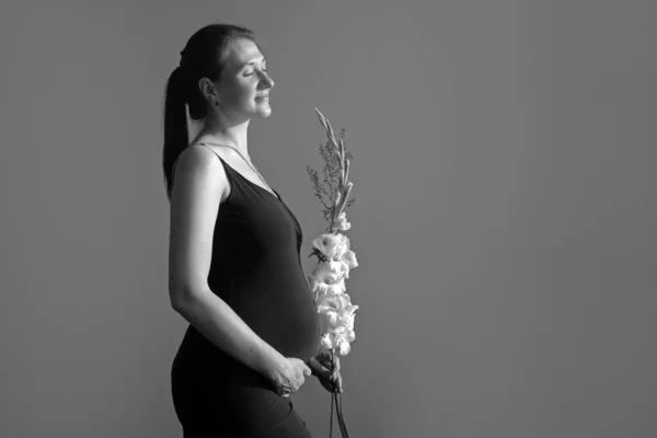 Vista laterale donna incinta con fiore tiene le mani sulla pancia foto monocromatica. Giovane donna in abito in attesa del parto. Festa della mamma Concetto di vacanza con spazio di copia. — Foto Stock