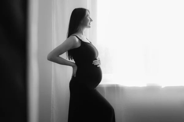 Silhouette di donna incinta in abito a casa dalla finestra toccare pancia. Foto in bianco e nero. — Foto Stock