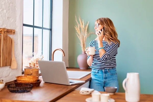 自宅のキッチンに立って窓の外を眺めながら ノートパソコンや携帯電話を使っている中年女性 内務省 — ストック写真