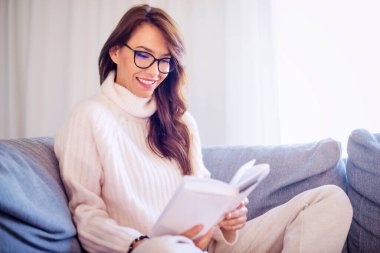 Evdeki kanepede rahat rahat oturup kitap okurken gülümseyen bir kadının fotoğrafı..