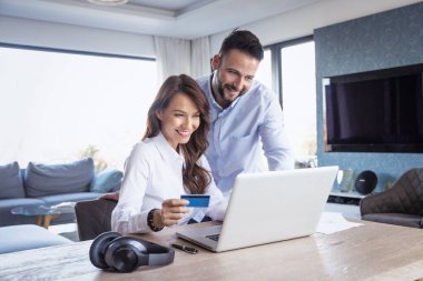 Evden online alışveriş. Elinde banka kartı tutan çekici bir kadın ve yanında gülümseyen bir adam dururken dizüstü bilgisayar kullanıyor ve internetten alışveriş yapıyor..  