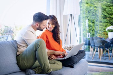 Mutlu çift kanepede birlikte dinleniyor. Neşeli kadın ve yakışıklı adam internette gezinirken dizüstü bilgisayar kullanıyor..