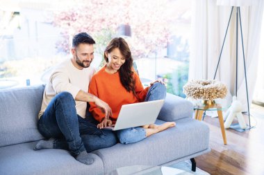 Mutlu çift kanepede birlikte dinleniyor. Neşeli kadın ve yakışıklı adam internette gezinirken dizüstü bilgisayar kullanıyor..