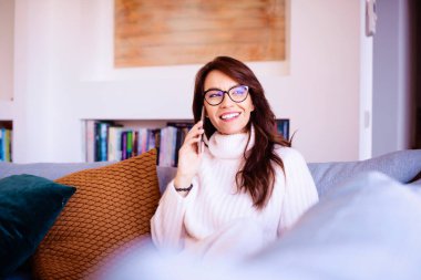 Çekici ve mutlu kadın akıllı telefonunu kullanıyor ve evdeki kanepede dinlenirken biriyle konuşuyor.. 