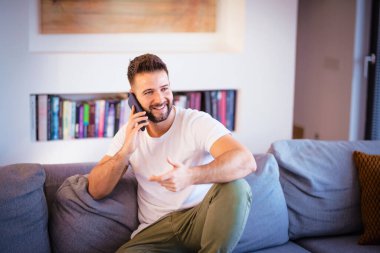 Shot ot handsome man relaxing on the sofa at home while talking with somebody on his smartphone.