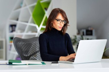 Kendine güveni tam orta yaşlı işkadını boğazlı kazak giyerken ofis masasında oturup yeni bir proje üzerinde çalışıyor.. 
