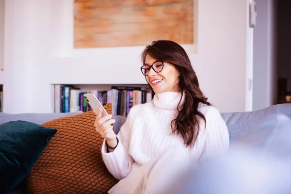 拍了一个迷人而快乐的女人使用她的智能手机和短信 同时在家里的沙发上放松 — 图库照片