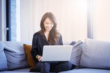 Mutlu bir kadının evdeki kanepede dinlenirken bilgisayarını kullanırken çekilmiş uzun bir resmi..