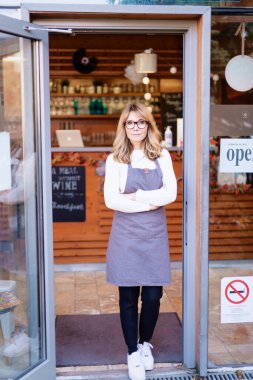 Gülümseyen kafe sahibi iş kadınının kollarını kavuşturup misafirini beklediği uzun bir sahne. Kapıda açık tabela.