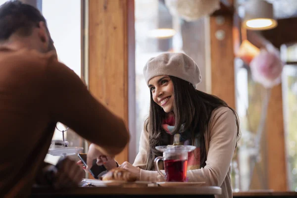 与一个难以辨认的男人坐在桌旁喝茶 头戴贝雷帽 头戴围巾的漂亮女人在咖啡馆里被拍了照 — 图库照片