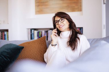 Çekici ve mutlu kadın akıllı telefonunu kullanıyor ve evdeki kanepede dinlenirken biriyle konuşuyor.. 