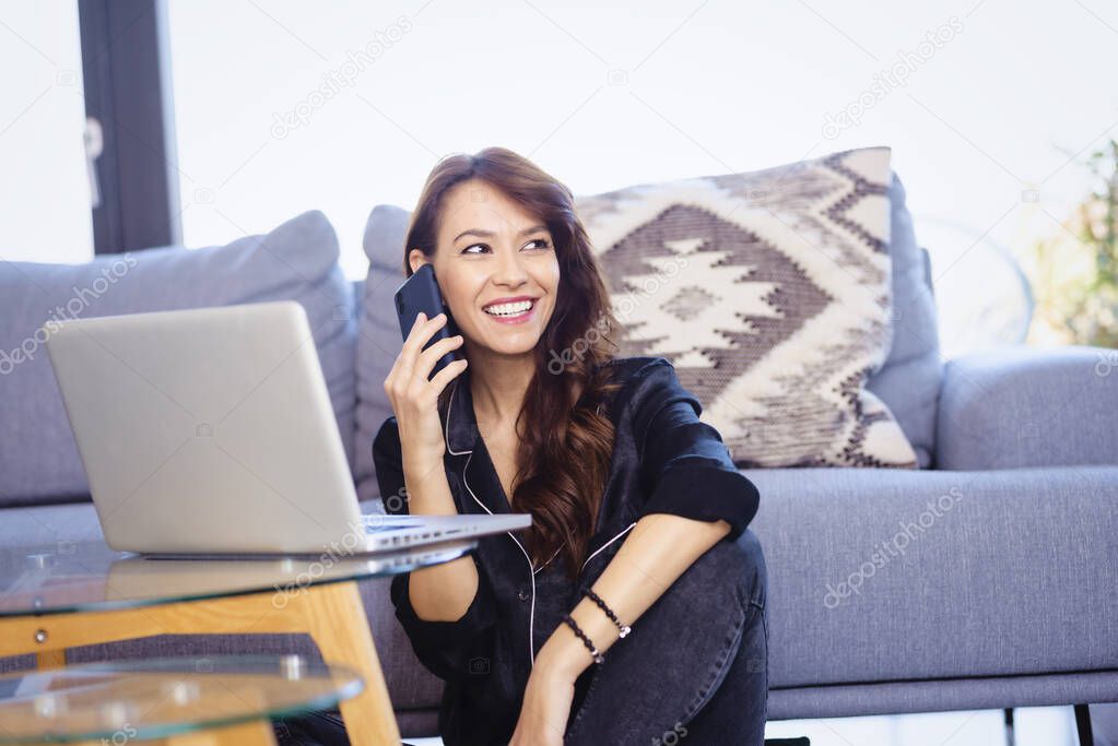 Cheerful attractive woman using mobile phone and laptop while working from home. Home office.