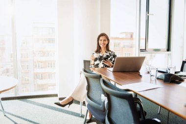 Full length shot of elegant businesswoman drinking her morning coffee and text messaging while sitting at office desk in the morning. 