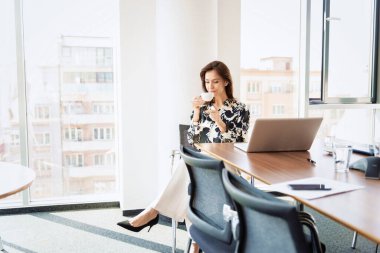 Full length shot of elegant businesswoman drinking her morning coffee while sitting at office desk in the morning. 