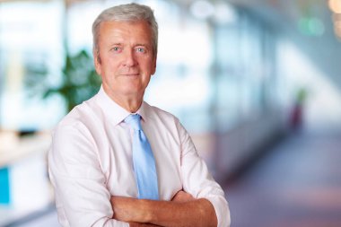 Portrait shot of executive senior businessman standing with arms crossed on the office corridor.