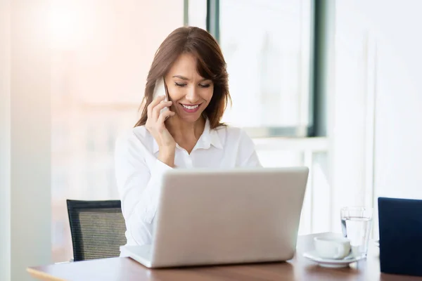 Smiling Businesswoman Talking Somebody Her Mobile Phone Using Laptop While — стоковое фото