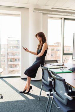 Full length shot of attractive and elegant businesswoman using her mobile phone while standing at the office.