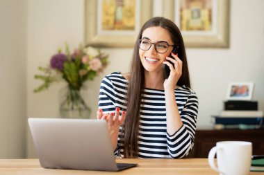 Happy young woman having a phone call and using laptop while sitting at desk at home. Home office.