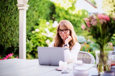Attractive middle aged woman using laptop while sitting at the backyard at home and working.