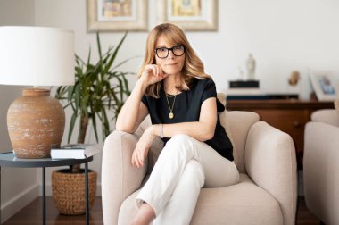 Portrait of confident middle aged woman looking deep in thought while relaxing at the armchair at home. 