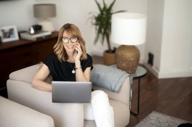 Confident woman using her laptop and mobile phone while sitting at the armchair and working from home. Home office.