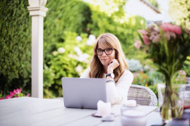Attractive middle aged woman using laptop while sitting at the backyard at home and working.