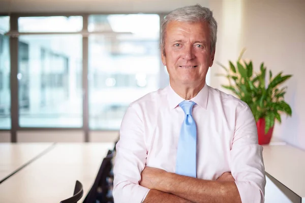 Executive senior businessman standing with arms crossed in a boardroom.