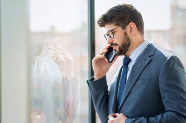 Handsome businessman wearing suit and using a mobile phone while standing at the office by the window.