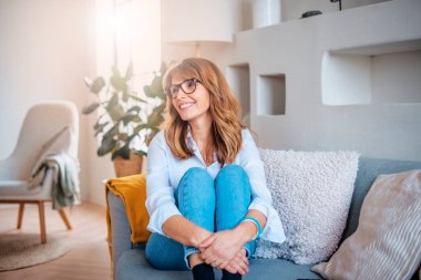 Candid shot of attractive middle aged woman looking away and smiling while relaxing on the sofa at home. 