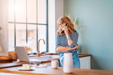 Confident middle aged woman talking with somebody on her mobile phone while standing at home in the kitchen.