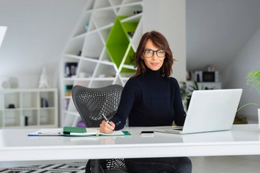 Kendine güveni tam orta yaşlı işkadını boğazlı kazak giyerken ofis masasında oturup yeni bir proje üzerinde çalışıyor.. 