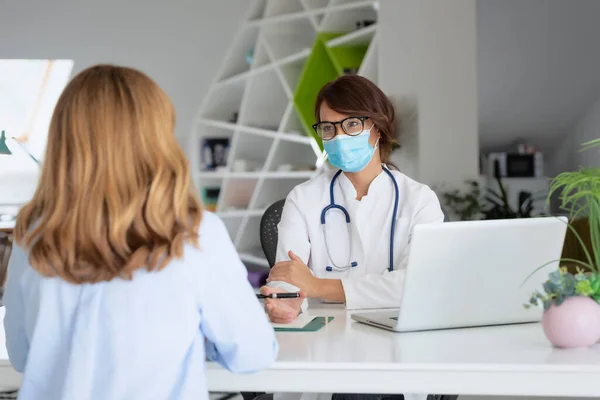 Female doctor wearing face mask while working in the office and listening to the patient.