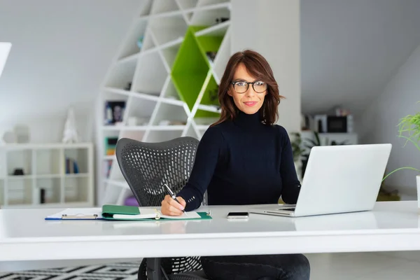 彼女のラップトップの後ろにオフィスデスクに座って 新しいプロジェクトに取り組んでいる間 タートルネックセーターを着て自信を持って中年のビジネスマンの女性 — ストック写真