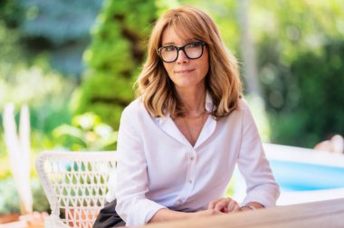 Close-up portrait shot of cheerful middle aged woman wearing glasses and white shirt while relaxing on the patio at home.