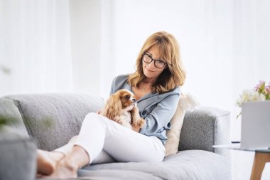 Middle aged woman relaxing on sofa at home with her cute spaniel puppy. 