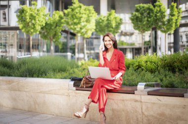 Parktaki bankta otururken bilgisayarını ve cep telefonunu kullanan çekici bir iş kadını. Kırmızı elbiseli, gülümseyen kadın..