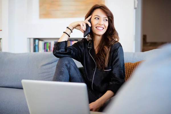 用笔记本电脑拍到了一个快乐的黑发女人坐在沙发上在家里工作的时候打电话 总部办公室 — 图库照片