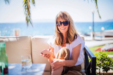 Deniz kenarındaki kafede otururken akıllı telefon ve kısa mesaj kullanan orta yaşlı bir kadın..