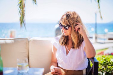 Orta yaşlı kadın düşünceli görünüyor ve deniz kenarındaki kafede otururken akıllı telefon kullanıyor..