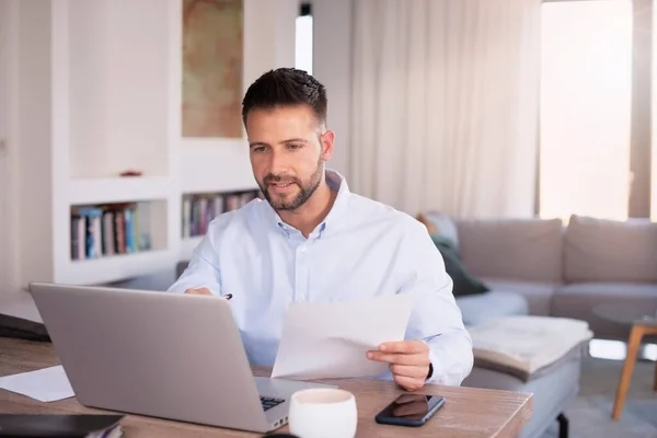 Middle aged businessman using laptop and doing some paperwork while working at home. Professional man holding document in his hand. Home office.