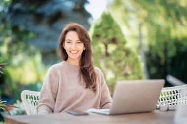 Balkonda dizüstü bilgisayarı olan kendinden emin orta yaşlı bir kadın oturuyor. Çekici kadın süveter giyiyor ve gülümsüyor..