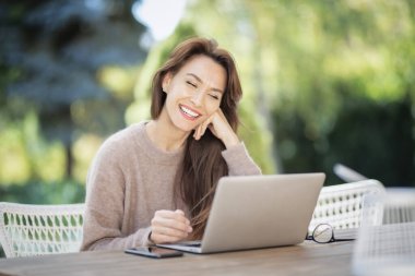 Kendine güvenen orta yaşlı bir kadın dizüstü bilgisayar kullanıyor ve balkonda otururken video görüşmesi yapıyor. Çekici kadın süveter giyiyor ve gülümsüyor..